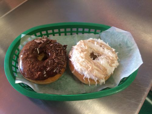 A green basket holds two donuts: one topped with chocolate and chocolate shavings, the other with white icing and coconut.