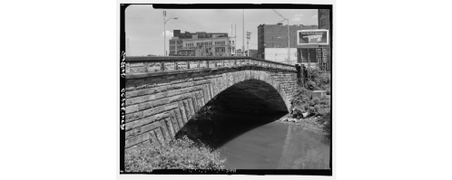 A stone arch bridge over a calm river, with buildings and a billboard in the background under a clear sky.