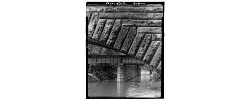 Black and white image of a stone bridge arching over a river, with a metal bridge visible in the background.