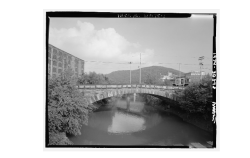A stone bridge spans a calm river, with trees and a factory building in the background under a cloudy sky.