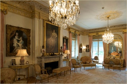 Elegant room with chandeliers, ornate decor, paintings, and vintage furniture, featuring a fireplace and large windows.