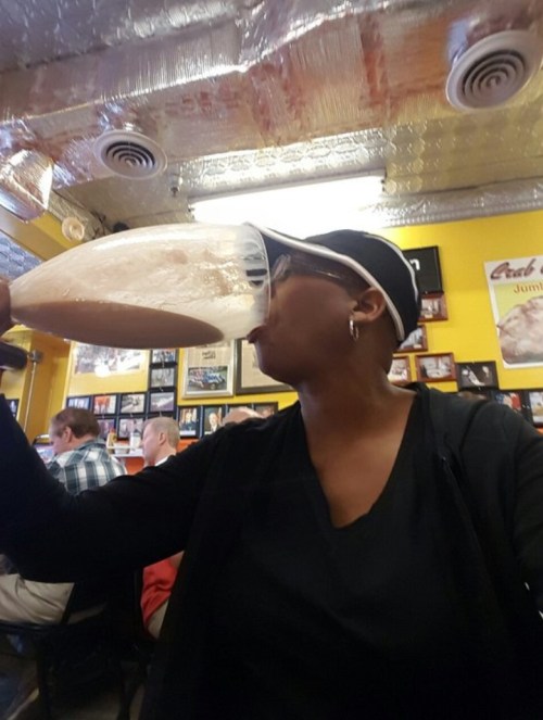 A person wearing a cap drinks from a large glass in a lively restaurant setting.