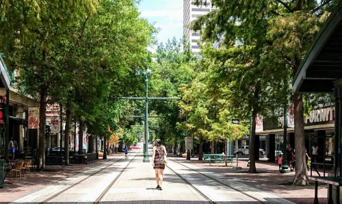 A woman walks down a tree-lined street with shops on either side and tram tracks in the center.