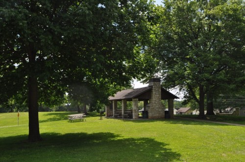 A stone picnic shelter surrounded by lush green grass and trees on a sunny day. Picnic tables are nearby.