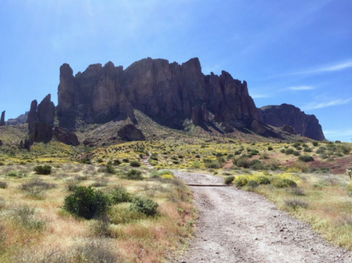 A dirt path leads through a grassy area towards a large, rocky mountain under a clear blue sky.
