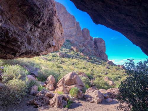 View from a cave opening, showcasing rocky terrain and lush greenery under a bright blue sky.