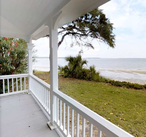 View from a porch overlooking a calm bay, with greenery and a sandy shore under a partly cloudy sky.