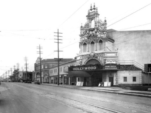 Historic black-and-white photo of a Hollywood theater and street, featuring ornate architecture and vintage cars.