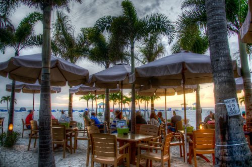 Beachside dining under palm trees with umbrellas, as the sun sets in the background. Relaxed atmosphere with patrons enjoying.