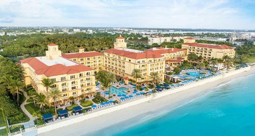 Aerial view of a beachfront hotel with red-tiled roofs, palm trees, and a pool area beside clear blue water.