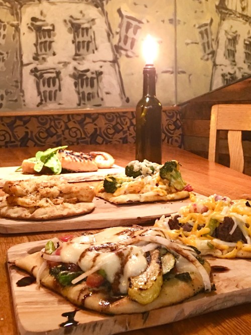 A wooden table with various flatbreads topped with vegetables and cheese, a candle, and a bottle in the background.