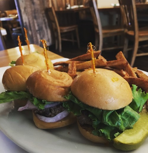 Three mini burgers with lettuce and onion, served with a side of crispy fries on a white plate.