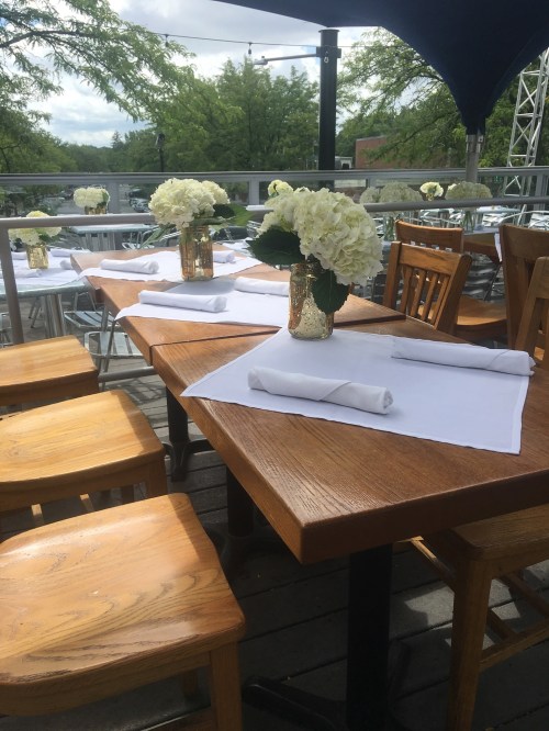 Outdoor dining setup with wooden tables, white tablecloths, and floral centerpieces under a shaded canopy.