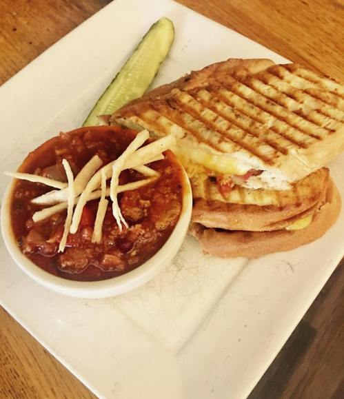 A grilled sandwich with melted cheese, served with a bowl of chili topped with crispy strips and a pickle on the side.