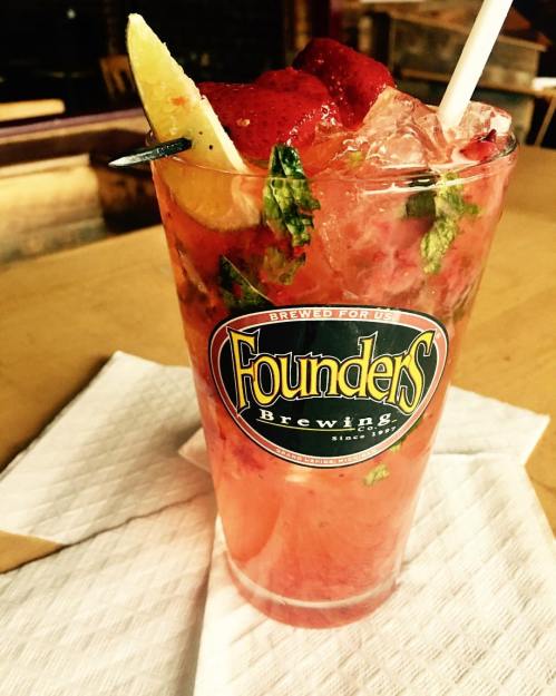 A refreshing cocktail in a Founders Brewing glass, garnished with mint, lemon, and strawberries, served over ice.
