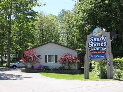 A welcoming campground entrance with a sign for Sandy Shores Campground, surrounded by trees and colorful flowers.