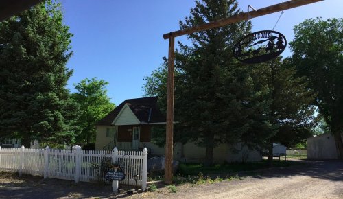 A quaint house with a sign hanging above, surrounded by trees and a white picket fence on a sunny day.