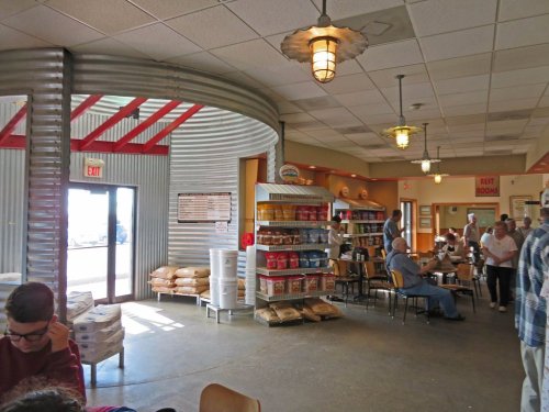 Interior of a casual dining space with tables, shelves of products, and a large entrance. People are seated and chatting.