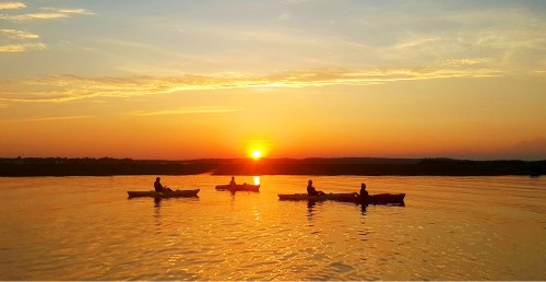 Silhouettes of four kayakers on a calm lake at sunset, with vibrant orange and yellow hues in the sky.