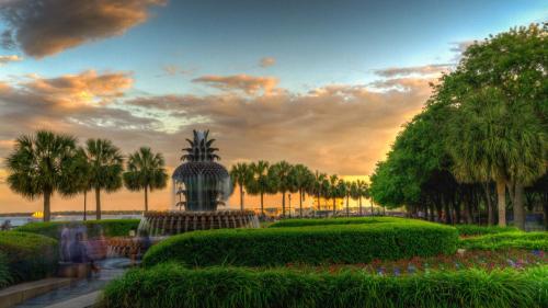 A scenic park at sunset featuring a fountain, palm trees, and colorful flowers under a cloudy sky.