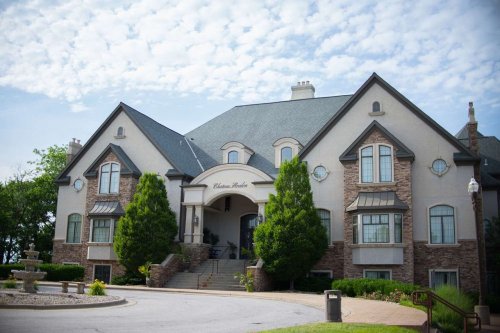 A large, elegant mansion with a stone facade, surrounded by greenery and a clear blue sky.