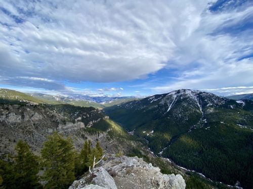 A panoramic view of mountains and valleys under a partly cloudy sky, with lush green forests and rocky terrain.