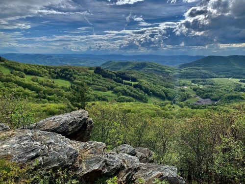 A panoramic view of lush green hills and valleys under a cloudy sky, with rocky foreground and distant mountains.