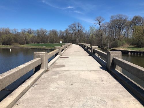 A concrete bridge stretches over a calm waterway, surrounded by trees and greenery under a clear blue sky.