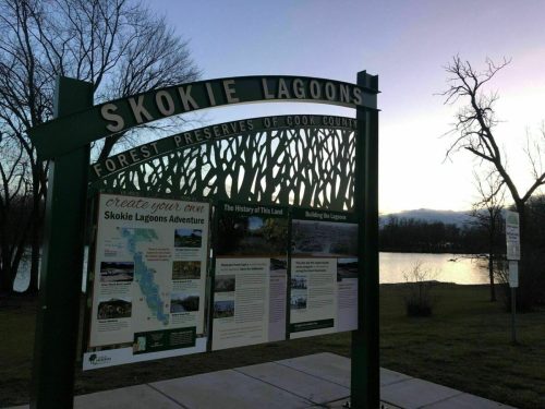 Sign for Skokie Lagoons with information panels, surrounded by trees and a view of the water at sunset.