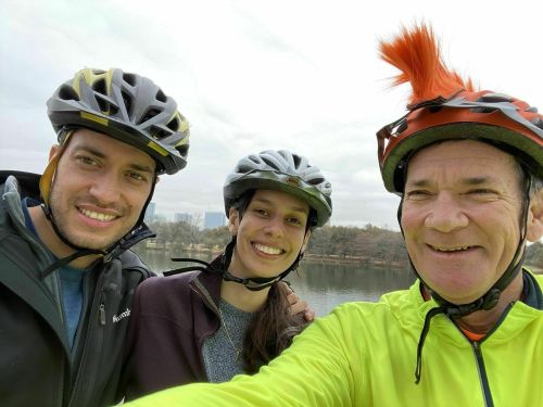 Three cyclists pose for a selfie by a lake, wearing helmets and smiling, with a city skyline in the background.