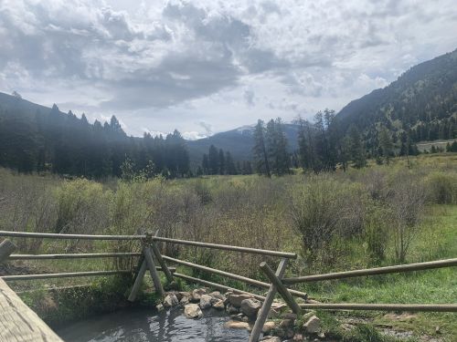 A serene landscape featuring a stream, wooden fence, and mountains under a cloudy sky, surrounded by lush greenery.
