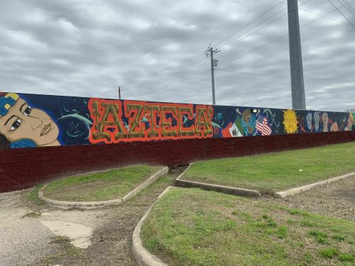 Colorful mural featuring the word "Azteca" and various cultural symbols on a wall, with a cloudy sky above.