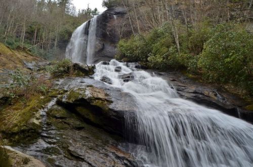 A cascading waterfall flows over rocky terrain, surrounded by lush greenery and trees in a serene natural setting.