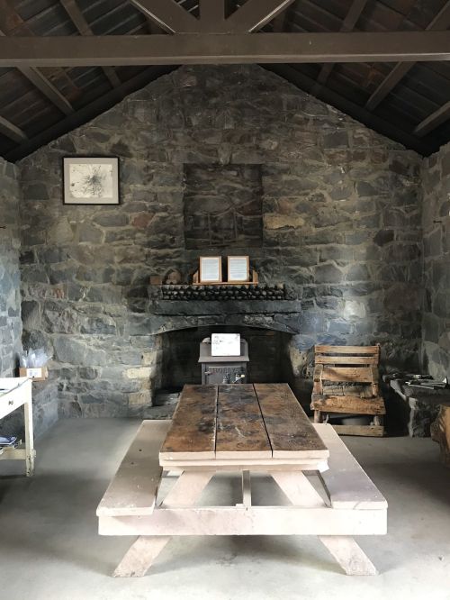 Interior of a rustic stone building featuring a wooden table, chairs, and a fireplace with a framed picture on the wall.