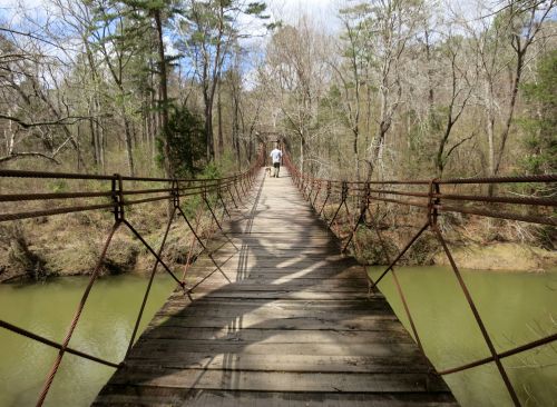 A person walks across a rustic suspension bridge over a calm, green river, surrounded by trees and a clear sky.
