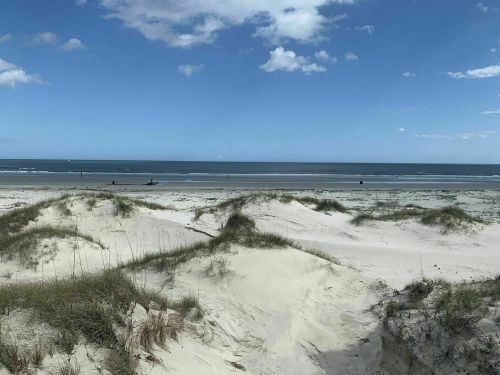 A serene beach scene with sandy dunes, grass, and a calm ocean under a blue sky with scattered clouds.