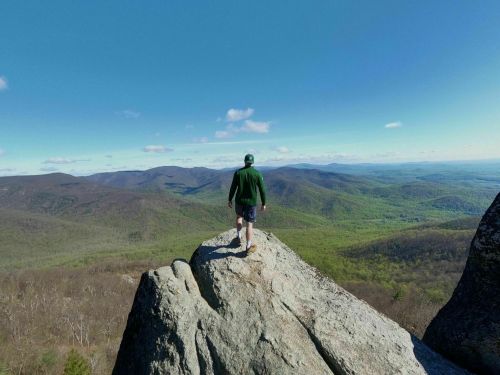 A person stands on a rocky outcrop, overlooking a vast landscape of green mountains and blue skies.