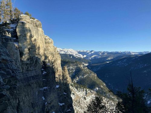 A panoramic view of snow-capped mountains and a rocky cliff under a clear blue sky.