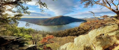 A panoramic view of a river winding through a valley, surrounded by autumn foliage and distant mountains under a blue sky.