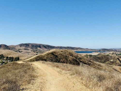 A dirt path winds through dry hills, leading to a lake in the distance under a clear blue sky.