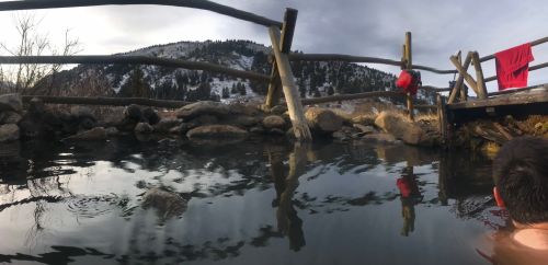 A serene hot spring surrounded by rocks and wooden fencing, with mountains in the background and a cloudy sky.