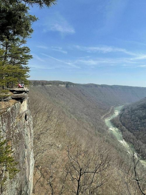 A person sits on a cliff edge overlooking a winding river and a valley of bare trees under a clear blue sky.