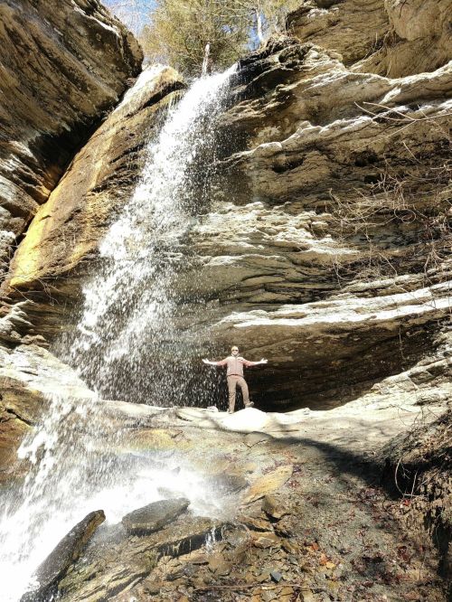 A person stands beneath a waterfall, arms outstretched, surrounded by rocky cliffs and flowing water.