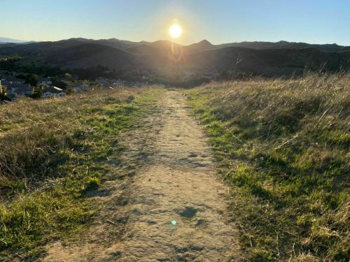 A sun sets over a grassy path leading through hills, with a view of a small town in the valley below.