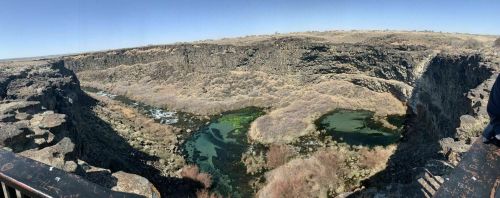 Panoramic view of a rocky canyon with two greenish ponds and sparse vegetation under a clear blue sky.