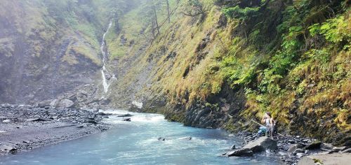 A serene river flows beside a rocky shore, with a waterfall cascading down a lush, green hillside in the background.