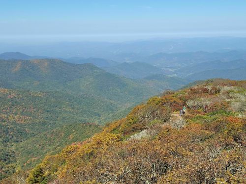 A panoramic view of rolling mountains with autumn foliage under a clear blue sky. A hiker is visible on the ridge.