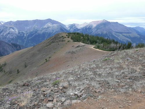 A scenic mountain landscape with rolling hills, rocky terrain, and distant peaks under a cloudy sky.