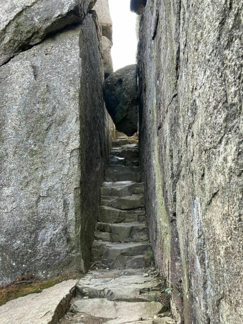 Narrow stone staircase between two large rock formations, leading upward with a boulder perched above.