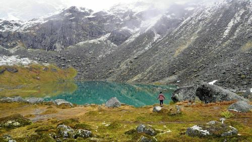 A person stands by a turquoise lake surrounded by rocky mountains and mist, with patches of snow and green grass.